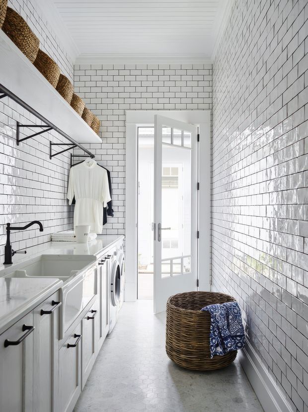 Bright, narrow laundry room with white glazed subway-tile walls, white painted cabinetry, a long utility bench with sink, front-loading washing machine, open shelving with woven baskets, hanging clothes, and a large wicker laundry basket. A glazed white door leads to a brighter adjoining space. The room has a clean, classic coastal feel with black hardware and plumbing accents.