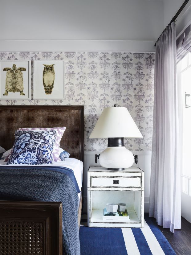 A bright, traditionally styled bedroom corner featuring a dark woven cane headboard and bed frame, navy quilted bedding, a patterned blue-and-white accent pillow, and a white bedside cabinet with glass-front storage and decorative trim. A large black-and-white floral patterned wallpaper covers the wall, with framed botanical and animal artwork above the bed. A white ceramic table lamp sits on the nightstand beside sheer lavender-grey curtains and a tall window. A navy-and-white striped rug adds contrast to the dark timber floor.