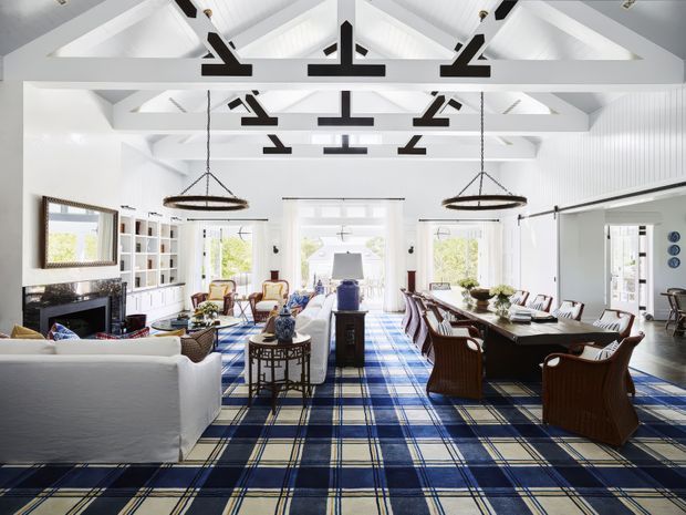 Bright, expansive open-plan interior combining a living room and dining area beneath a high white timber-trussed ceiling. The room features white panelled walls, exposed dark structural beams, large windows and glazed doors, built-in shelving, a fireplace, a long dining table, and a bold navy-and-cream plaid carpet. Dark wood furniture and woven pendant lights add contrast.