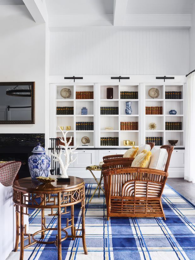 A bright, refined living room with white built-in display cabinetry, vertical wall panelling, exposed ceiling beams, a fireplace, and extensive shelving filled with books and decorative objects. Woven rattan and bamboo-style armchairs, a round wood-and-wicker side table, blue-and-white ceramic decor, and a large blue plaid rug create a traditional coastal-inspired interior.