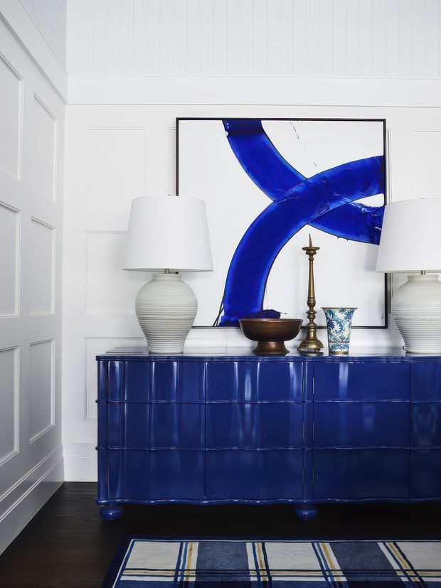 A refined interior vignette featuring a glossy deep-blue tiled sideboard against white decorative wall panelling. The sideboard is styled with two white ceramic table lamps, a brass candlestick, a dark bowl, and a blue-and-white patterned vessel, with a large abstract artwork in blue and white above. Dark timber flooring and a blue, cream, and grey checked rug complete the high-contrast composition.