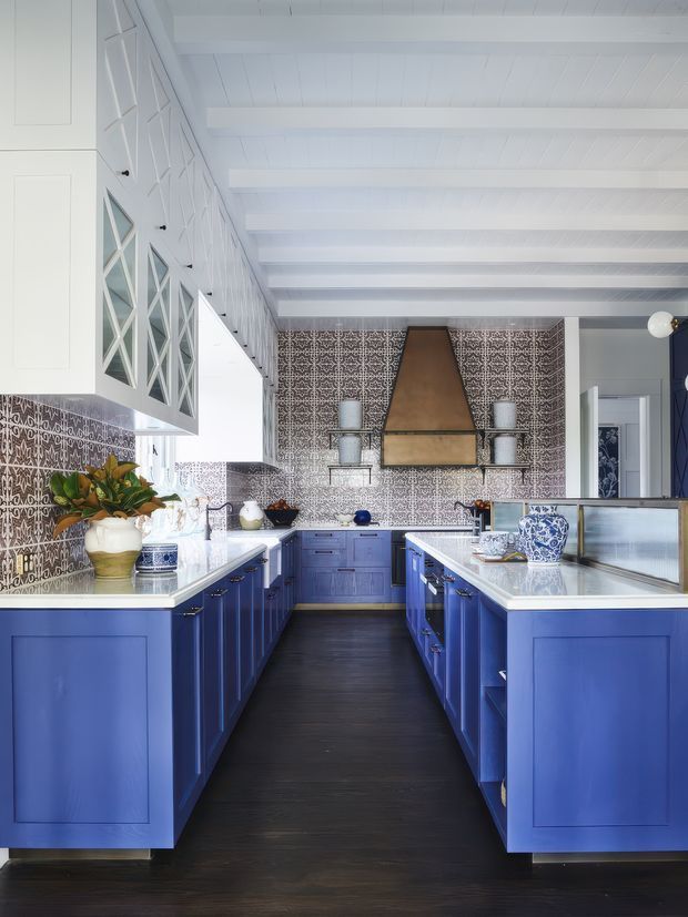 A bright, galley-style kitchen with vivid blue shaker cabinetry on both sides, white countertops, and a dark timber floor. The room features extensive patterned tile splashbacks, a large bronze-toned rangehood, open shelving, glass-front upper cabinets, white painted ceiling beams, and decorative blue-and-white ceramics. The overall look is colourful, traditional coastal-inspired, and highly detailed.