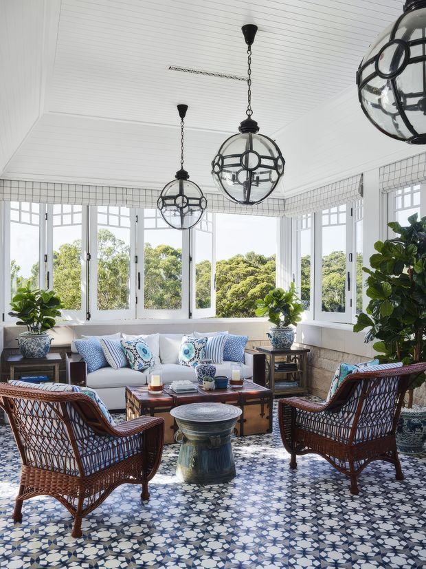 Bright enclosed sunroom filled with natural light, featuring wraparound white-framed windows, a white beadboard ceiling, patterned blue-and-white tiled floor, and a relaxed seating arrangement of a white sofa, wicker armchairs, and a wooden coffee table. Spherical black metal pendant lights and abundant potted greenery add character, while the windows overlook leafy trees.