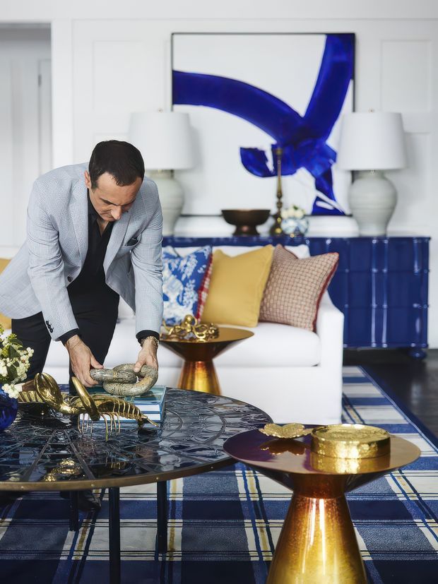 A boldly styled living room with a white sofa, patterned blue-and-white rug, navy built-in cabinetry, gold-toned sculptural coffee tables, white lamps, and a large abstract artwork with vivid cobalt-blue forms. A person is arranging decorative objects on the glass coffee table.