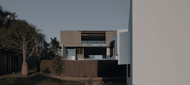 Exterior view of a contemporary multi-level residence, largely obscured by a tall light-coloured foreground wall. The house features a rectilinear form, dark and timber-toned facade sections, broad recessed openings, horizontal timber screening, and a flat roofline. Landscaping, a boundary fence, and mature trees frame the approach.
