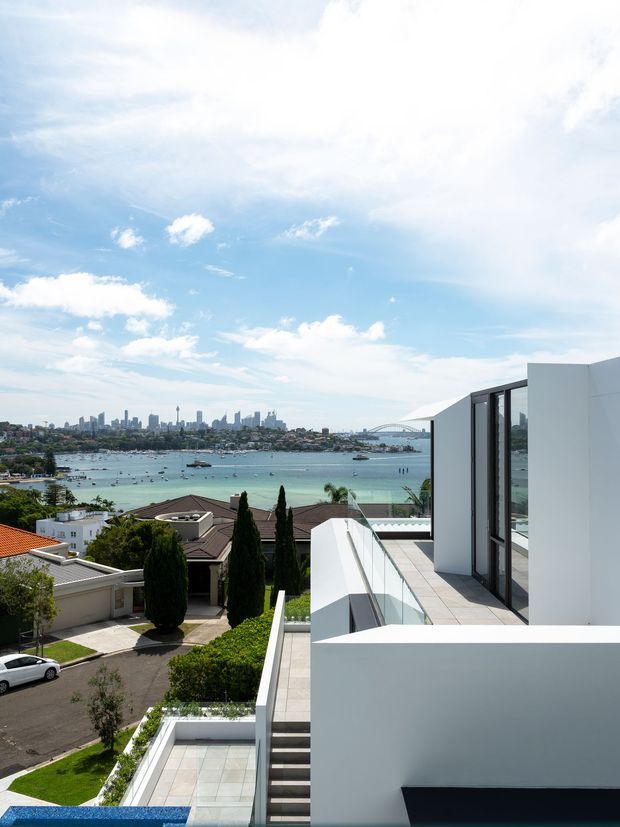 A bright contemporary architectural exterior overlooking a calm harbour with a city skyline and bridge in the distance. The white multi-level residence features clean geometric forms, expansive dark-framed glazing, tiled terraces, glass balustrades, and landscaped gardens with tall slender trees. The view includes neighbouring homes, water, boats, and urban surroundings.