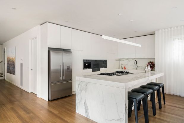 Bright, open-plan contemporary kitchen with extensive white flat-panel cabinetry, a large marble-look waterfall island with integrated cooktop and seating for four, stainless-steel French-door refrigerator, built-in black appliances, and a marble-look splashback. Warm timber flooring and sheer curtains soften the predominantly white scheme.