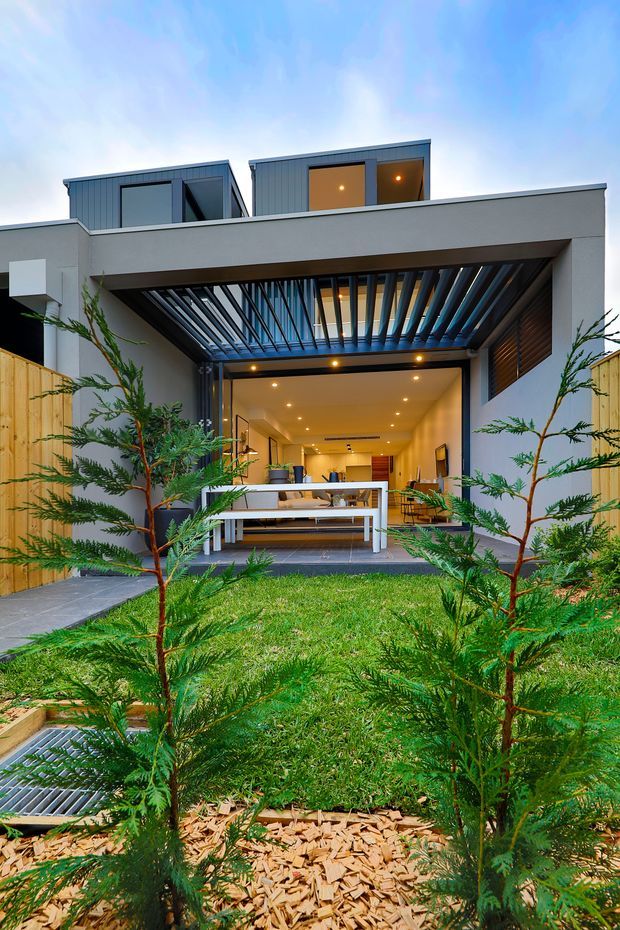 Exterior view of a contemporary multi-level residence with a covered outdoor entertaining area and adjustable dark louvre pergola over a glazed opening. The open living space beyond contains seating and a kitchen or dining zone, while a landscaped lawn, young trees, timber fencing, and paved paths occupy the foreground.