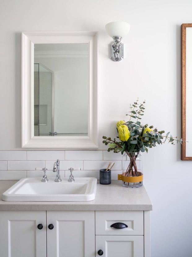 Bright, refined bathroom vanity area with a white framed mirror, chrome wall sconce and traditional three-piece tapware above a white inset basin. The vanity has a pale stone-look benchtop and white shaker-style cabinetry with dark pulls. White horizontal subway tiles form the splashback, complemented by a floral arrangement in a glass vase.