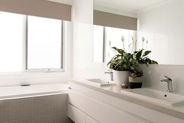 Bright, clean contemporary bathroom with a long white vanity, integrated double basins and chrome tapware. A large wall mirror reflects the room, while a freestanding bathtub sits beneath a window with taupe roller blinds. Neutral white and light-grey finishes create a calm, minimalist atmosphere, with a potted plant adding greenery.