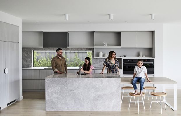 Bright, contemporary family kitchen with a large pale stone island, integrated grey cabinetry, open shelving, built-in appliances, and a connected timber dining table. Family members gather around the island and dining area. The space has a clean, restrained palette and generous natural light.