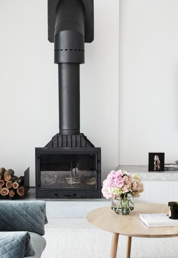 A bright contemporary living room centred around a black freestanding wood-burning fireplace with a prominent vertical flue. The room features pale walls and flooring, a light grey-blue quilted sofa, a round light timber coffee table, a white built-in cabinet, and a vase of soft pink and cream flowers.