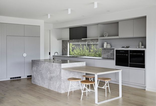 Bright contemporary kitchen with a large grey stone island, integrated sink and tap, pale timber flooring, grey built-in cabinetry, open shelving, black appliances, and a white-framed dining table with timber stools. A wide window above the rear benchtop brings in greenery and natural light.