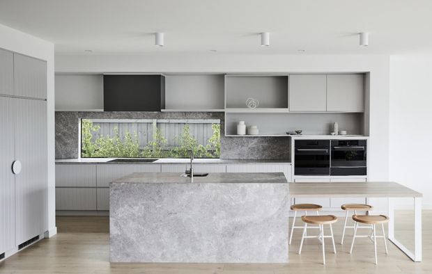A bright contemporary kitchen with a large grey stone island, integrated cabinetry, open shelving, built-in black appliances, and a light timber dining table with stools. Vertical window glazing brings greenery and a fenced outdoor view into the space. The palette is restrained in white, pale grey, and natural wood tones.