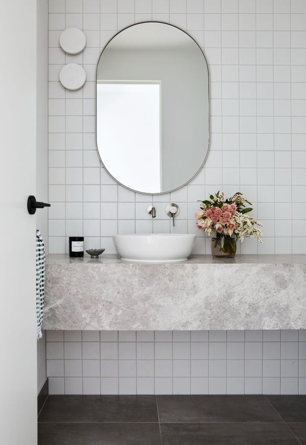A refined contemporary bathroom vanity with a large rounded-arch mirror, white vessel basin, wall-mounted brushed-metal tapware, and a substantial pale stone vanity slab. White square wall tiles with dark grout cover the wall, while large dark floor tiles provide contrast. Minimal black hardware and soft floral styling add understated accents.