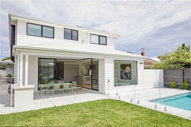Two-storey white weatherboard home viewed from the backyard, with large glazed living areas opening onto a paved terrace and a rectangular swimming pool. The exterior has a low-pitched roof, simple contemporary detailing, extensive aluminium-framed glazing, and a fenced suburban garden setting.