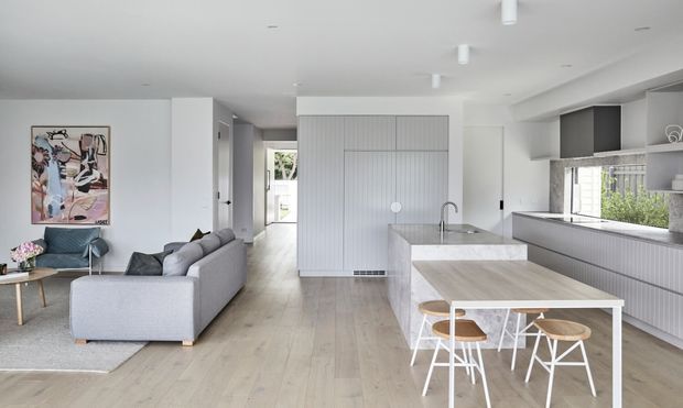 Open-plan contemporary kitchen, dining and living area with pale timber flooring, white vertical-panel cabinetry, a light stone island, grey sofa seating, and large windows. The bright, restrained interior includes a compact dining table and modern pendant-free ceiling lighting.