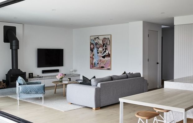 Contemporary open-plan living area viewed from the kitchen, with a grey sectional sofa, wall-mounted television, freestanding black wood-burning stove, light timber flooring, pale walls, and a dining or kitchen island with stools in the foreground. The room is softly furnished with a patterned blue armchair, round coffee table, artwork, and neutral finishes.