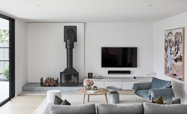 Bright contemporary living room with a large grey sectional sofa in the foreground, a low timber coffee table, sculptural lounge chairs, and a wall-mounted television. A freestanding black wood-burning stove with exposed flue forms a strong feature beside the built-in media cabinetry. Full-height glazing opens toward an outdoor area, while an abstract artwork adds colour to the otherwise neutral interior.