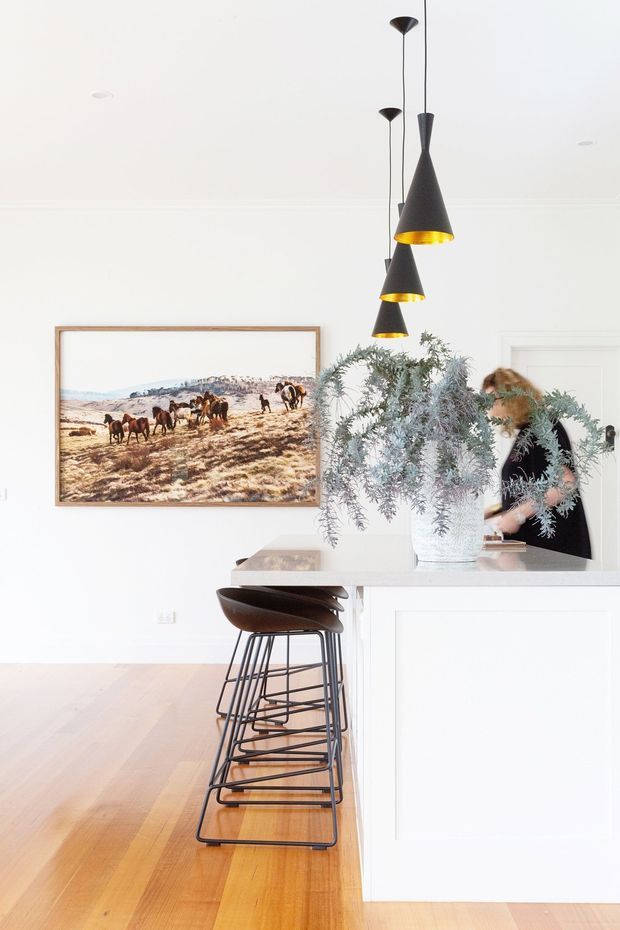 Bright, refined kitchen interior with a white island and raised breakfast bar, lined with dark sculptural bar stools. Three black cone pendant lights hang above the island, while a large framed landscape photograph and a vase of silvery foliage add visual interest. The room features pale timber flooring, white walls and cabinetry, and a clean, airy aesthetic.