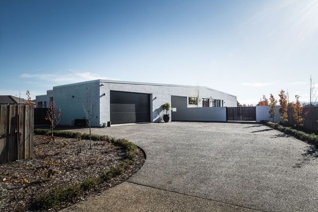 Single-storey contemporary residential building viewed from the front, with a low flat-roofed form, pale grey masonry or rendered facade, large dark garage door, enclosed front yard, and broad textured driveway. Sparse landscaping and strong sunlight create a clean, understated exterior composition.
