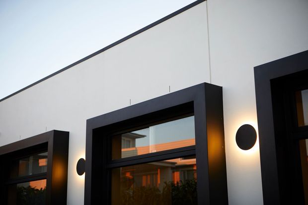 Close-up exterior view of a contemporary white rendered facade with bold black-framed windows and projecting rectangular surrounds. Round wall-mounted lights flank the openings, with warm interior and reflected sunset tones visible through the glazing.