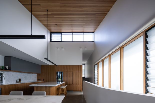 Contemporary open-plan kitchen and dining space with a long island, dark integrated cabinetry, timber wall panelling, a marble-look benchtop, and extensive timber-framed glazing. A high timber-lined ceiling and clerestory windows create an airy, naturally lit interior, while linear pendant lights and louvred windows add a refined modern character.