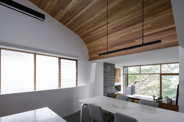 Contemporary open-plan dining and living interior with a large marble-look dining table, upholstered chairs, expansive windows, and a dramatic warm timber-clad vaulted ceiling. Linear black pendant lights and a dark tiled fireplace feature add contrast, while the elevated outlook through the glazing brings in natural light.