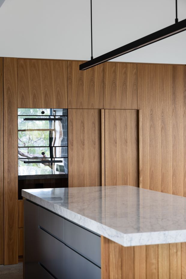 A refined contemporary kitchen featuring extensive vertical timber wall panelling and matching island cabinetry, a pale veined stone benchtop, dark matte drawer fronts, and a long black linear pendant light suspended from a high ceiling. A narrow glazed opening provides views to trees outside, while integrated timber doors and minimal detailing create a calm, streamlined composition.