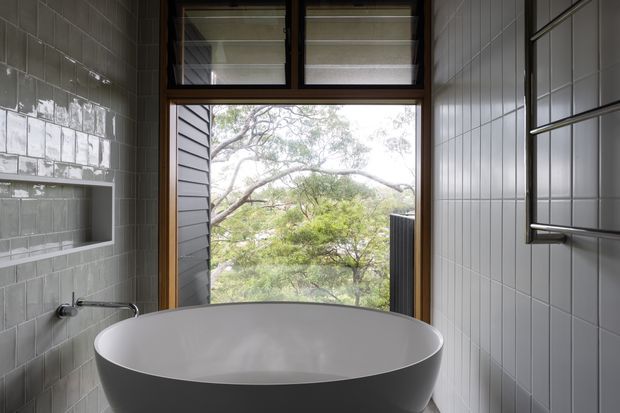 Compact contemporary bathroom featuring a freestanding oval bathtub positioned in front of a large timber-framed window. The walls are clad in small glossy grey rectangular tiles, with a recessed tiled niche and wall-mounted tapware on the left. Dark-framed louvre windows sit above the main glazing, and a towel rail is mounted on the right wall. Leafy trees are visible outside.