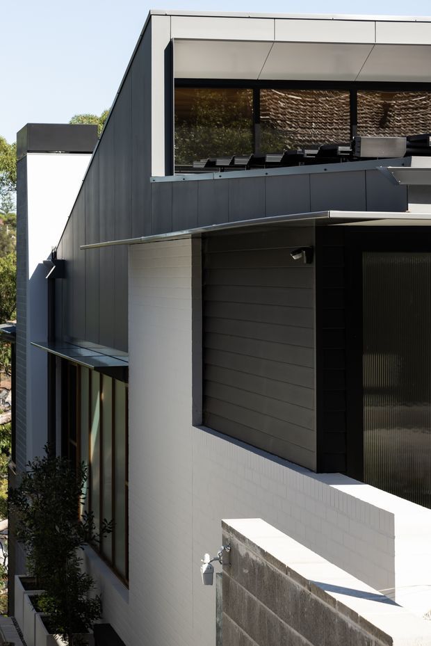 A close exterior view of a contemporary multi-level residence with strong geometric forms, dark vertical cladding, white tiled masonry, broad horizontal roof overhangs, and large upper-level glazing. A narrow planted side passage, garage or service openings, and a concrete block boundary wall are visible. The facade has a restrained black, white, and grey palette with pronounced sun and shadow contrast.