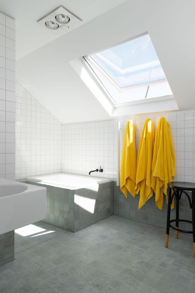 Bright, compact tiled bathroom with a white bathtub beneath a large skylight, a partial white basin in the foreground, and vivid yellow towels hanging on the wall. Pale blue-grey floor and lower wall tiles contrast with white square upper wall tiles. A black stool and dark tapware add accents, while recessed ceiling lights and strong daylight create an airy atmosphere.
