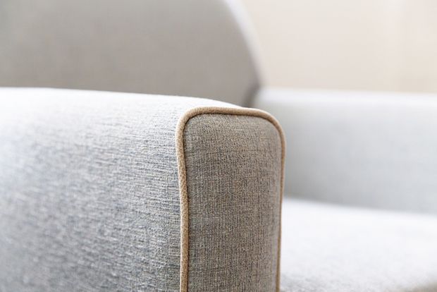 Close-up detail of a softly upholstered sofa or armchair armrest with rounded edges and contrasting beige piping. The surrounding upholstery is light grey, set against a bright neutral background with a minimalist, tactile appearance.