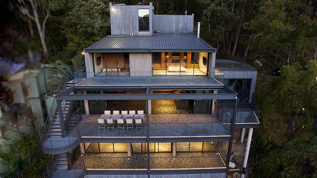 A multi-level contemporary house nestled among dense trees, viewed from above. The home features extensive outdoor decks and terraces with dark metal balustrades, a corrugated metal pitched roof, timber and dark-clad walls, large glazed openings, and warm interior lighting. Curved external stairs and elevated decks create a strongly vertical, multi-storey form integrated into a wooded site.