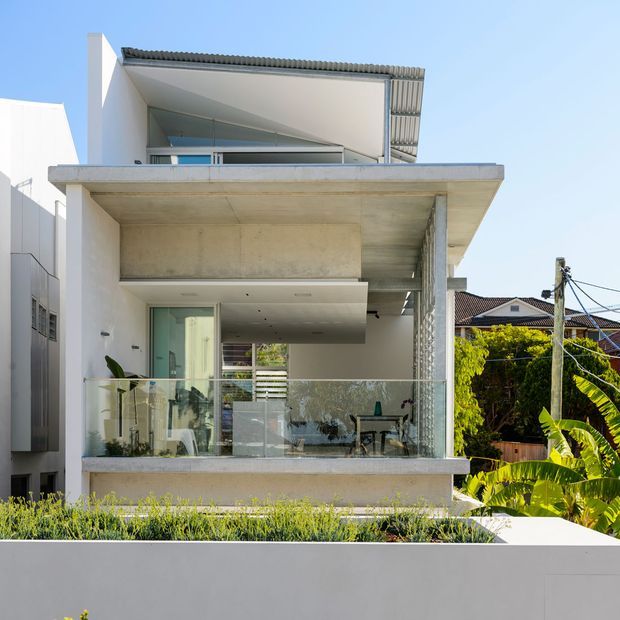 Front exterior view of a contemporary multi-level residence with exposed concrete balconies and slab edges, white rendered walls, extensive glazing, a corrugated metal roof, and transparent glass balustrades. Tropical planting and neighbouring suburban homes are visible around the property.
