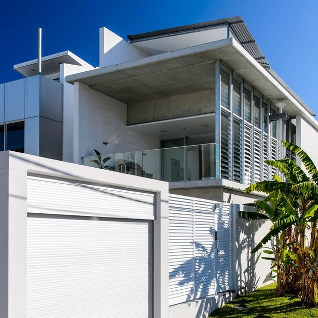 A bright contemporary multi-level residential facade with white rendered walls, prominent exposed concrete balcony soffit, glass balustrades, horizontal louvres, roller garage door, and corrugated metal roof elements. Tropical banana plants sit beside the building, while strong sunlight creates crisp shadows against a deep blue sky.