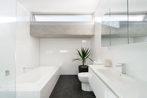 Bright contemporary bathroom with a freestanding white bathtub, wall-mounted white vanity and basin, toilet, large mirror, dark tiled floor, and a concrete feature ledge beneath high-level windows. A potted plant adds a natural accent.