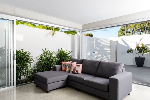 Bright contemporary indoor-outdoor living area with a large dark grey sectional sofa positioned beside expansive sliding glass doors. White walls, exposed concrete-style ceiling beams, pale flooring, tropical planting, and a private paved courtyard with louvred screening create a clean, airy connection to the outdoors.