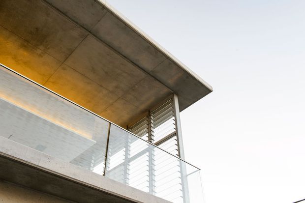 Close-up architectural exterior view of a modern concrete building, featuring a deep flat roof overhang, exposed concrete soffit and balcony slab, clear glass balustrading, and horizontal louvres providing solar shading. The composition emphasizes clean lines, raw materials, and contemporary passive shading elements against a bright sky.