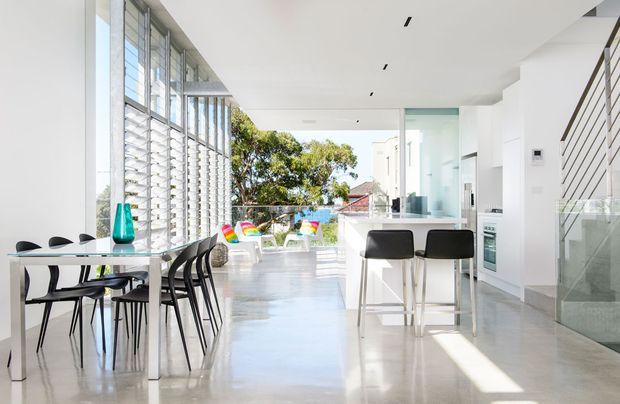 Bright, open-plan contemporary interior combining a dining area, kitchen island and stairway. The space features polished light-grey concrete flooring, white walls and ceiling, extensive floor-to-ceiling glazing with horizontal louvres, a white kitchen island with dark bar stools, a minimalist dining table and chairs, and a glass-sided staircase. Strong daylight creates an airy, clean atmosphere.