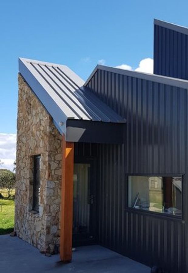 Exterior view of a compact modern building with intersecting pitched roof forms, dark vertically ribbed metal cladding, a natural irregular-stone wall, and a recessed entry beneath a projecting roof canopy. A small window and covered concrete entry area are visible, with blue sky and landscape in the background.