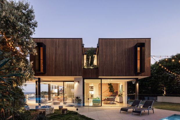 Twilight exterior view of a contemporary two-storey residence with two projecting dark timber-clad upper volumes, extensive floor-to-ceiling glazing at ground level, and a central recessed courtyard containing tall cacti. The home opens onto a paved terrace and landscaped outdoor area with lounge chairs, a pool edge, and warm festoon lighting.