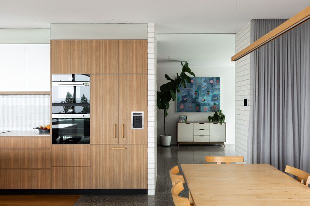 A bright contemporary open-plan kitchen and dining area. The kitchen features full-height warm timber cabinetry with integrated black appliances, white upper cabinetry, and a pale benchtop. A tiled structural column separates the kitchen from a dining space with a large natural timber table and matching chairs. Grey curtains, a grey tiled floor, indoor plants, and colourful artwork add contrast.