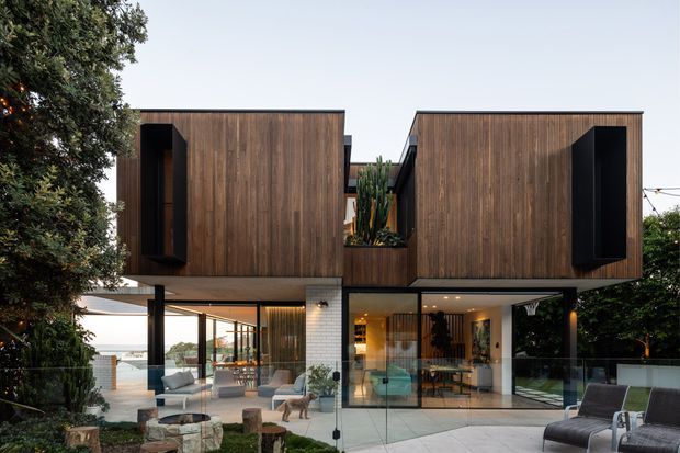 A contemporary two-storey residence with strongly defined box-like volumes clad in vertical dark timber, contrasting black window reveals, and extensive floor-to-ceiling glazing at ground level. The transparent lower floor opens onto landscaped outdoor areas and a patio, while a central recessed courtyard contains tall planting. The composition includes a flat roofline, cantilevered upper forms, and a warm, refined interior visible through the glazing.