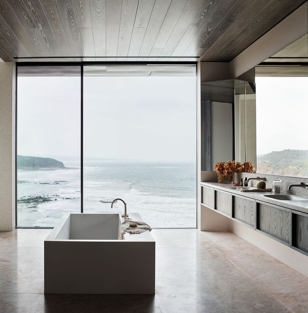A luxurious contemporary bathroom with a freestanding rectangular bathtub positioned before floor-to-ceiling glazing overlooking the ocean and rocky coastline. The room features a double vanity with integrated sinks, large mirrors, grey wood-grain cabinetry, a pale stone or concrete benchtop, dark slim window frames, and dramatic timber ceiling panelling. The palette is restrained in grey, taupe, white, and natural wood tones, creating a serene coastal atmosphere.