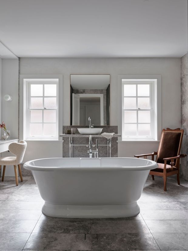 A refined, symmetrical bathroom interior centered on a freestanding white bathtub. Two white-trimmed windows flank a mirrored vanity with a vessel sink and polished metal tapware. Large grey stone-look floor tiles, grey wall finishes, a sculptural white chair, and a brown leather armchair create a calm, classic atmosphere.
