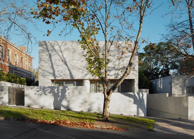 Street-facing view of a contemporary residential building in an established suburban setting. The house has a minimalist pale rendered facade, a simple box-like form, narrow vertical glazing or screening, and high solid boundary walls. Mature deciduous trees and neighboring period-style brick architecture frame the property.