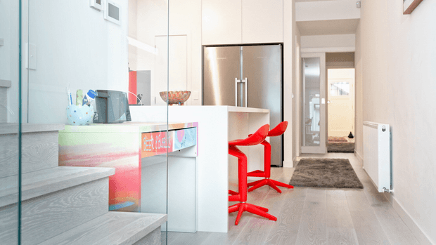 Bright contemporary open-plan kitchen viewed from a stairway, featuring a white island with colourful inset artwork, stainless-steel double-door refrigerator, two vivid red bar stools, pale timber-look flooring, pale cabinetry, and a glass stair balustrade. A hallway extends beyond the kitchen.