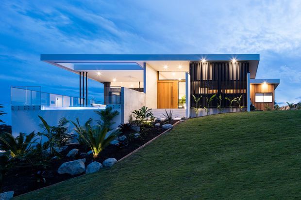 A contemporary single-storey residence photographed at dusk, featuring a broad flat roof with deep overhangs, expansive open glazing, warm timber entry doors, white rendered walls, and a prominent vertical black louvred screen. The home is set into a landscaped sloping site with tropical planting, stone edging, and a visible illuminated pool or water feature.