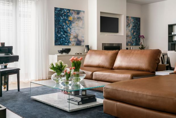 A refined contemporary living room featuring a large brown leather sectional sofa, glass coffee table with a marble-look lower shelf, dark area rug, white walls, sheer floor-to-ceiling curtains, abstract blue artwork, a wall-mounted television, and a recessed fireplace. A piano is visible near the window, with flowers and decorative objects adding warmth.